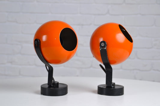 Two orange space-aged eyeball spherical wall lights on black stands against a light gray brick wall.