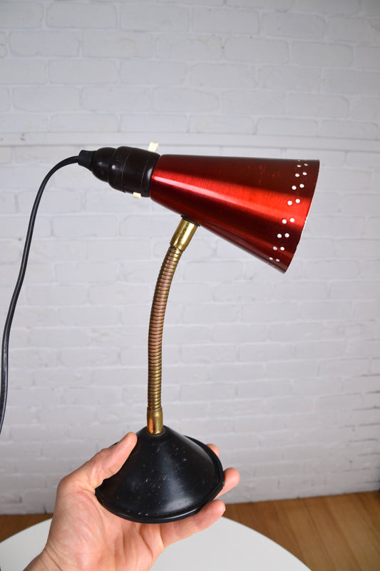 Hand holding a vintage-style desk lamp with a red shade against a white brick wall.